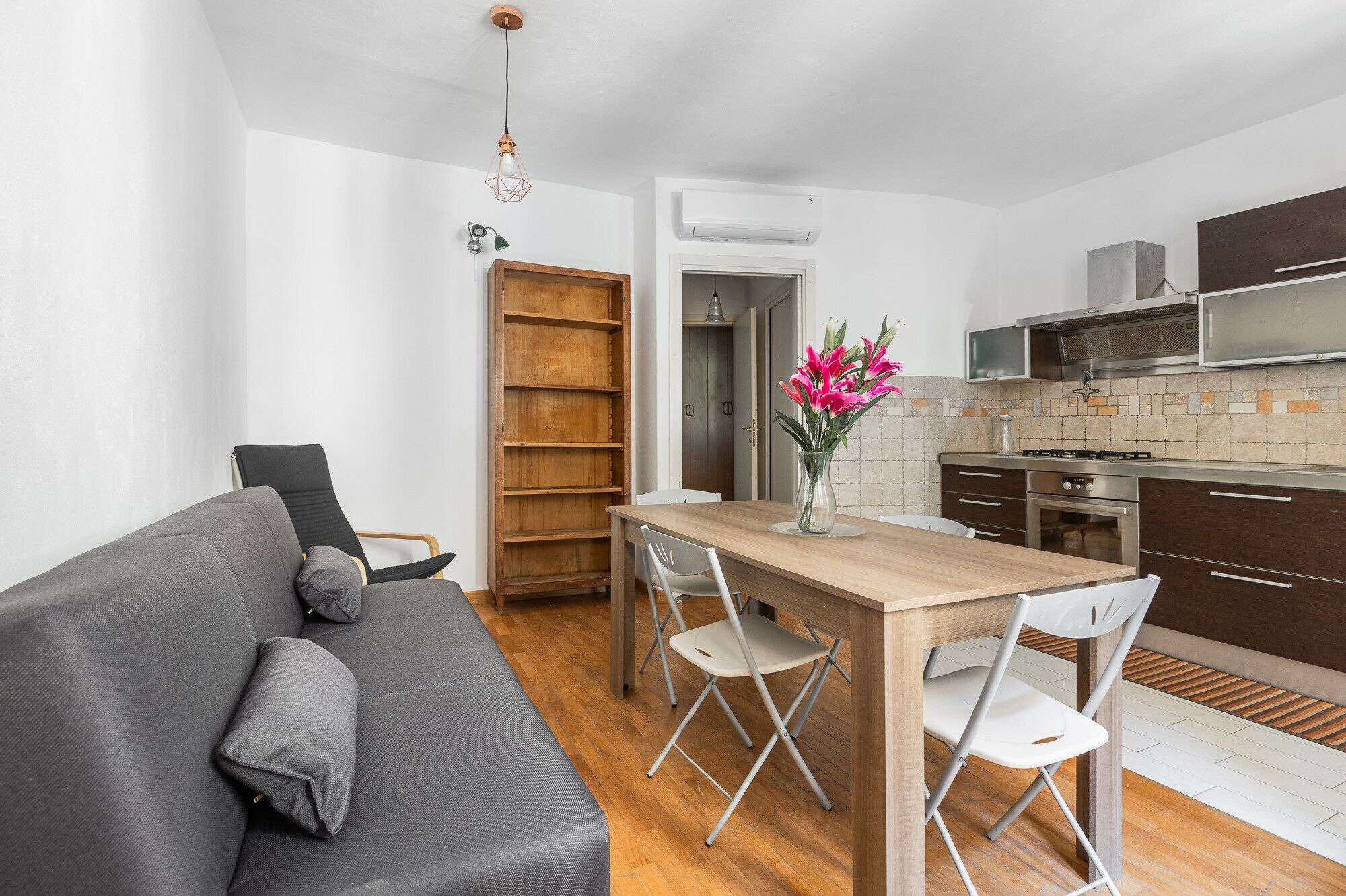 A compact living space with a sofa, dining table, chairs, a kitchen area with a stove, and a bookshelf.