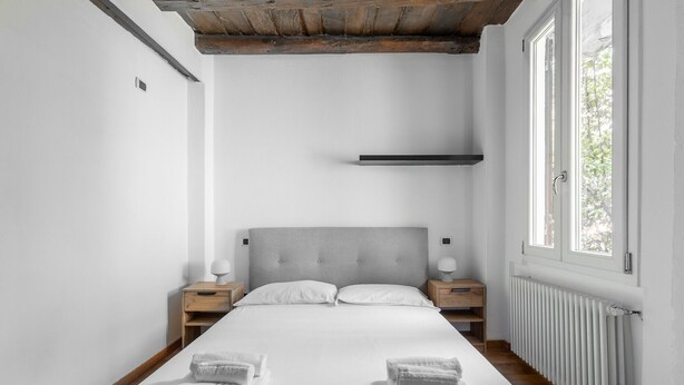 A bedroom with a bed, two bedside tables, a nightstand, a radiator, and a window. - Casa Centotrecento by Wonderful Italy (Bologna)