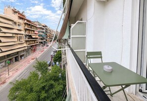 En balkon med et bord og en stol, der vender ud mod en gade med bygninger og parkerede biler.