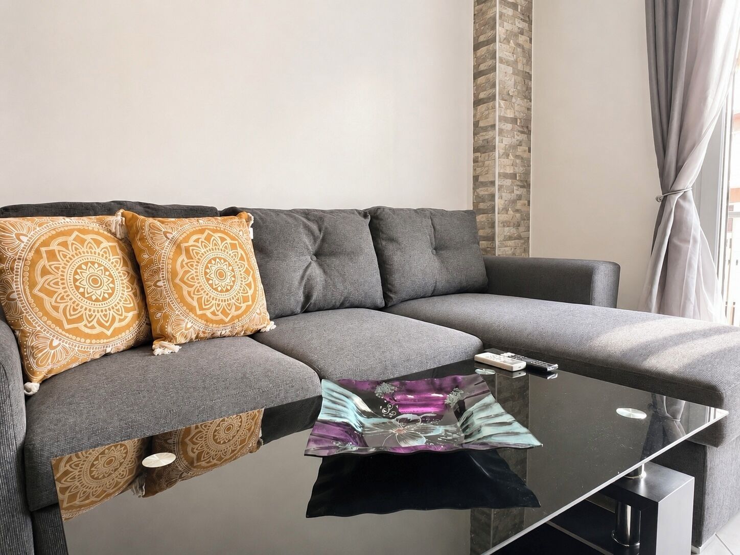 Un sofá gris con dos cojines estampados, una mesa de centro de cristal y una ventana con cortinas.