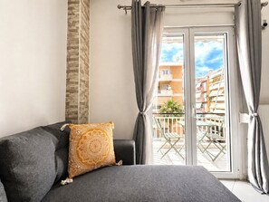 Une pièce avec un canapé gris, un coussin à motifs et un balcon offrant une vue sur des immeubles et un palmier.