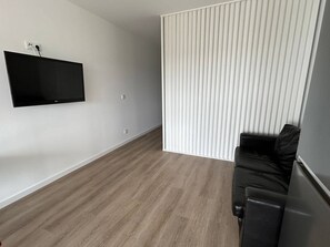 A room with a flat-screen TV mounted on the wall, a black leather sofa, and a wooden floor. - Apartamento T1 com Terraço (Coimbra)