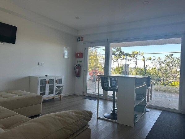 A modern room with a sofa, a small cabinet, a bar area, and a large sliding glass door leading to an outdoor area. - Apartamento T2 com Terraço (Coimbra)