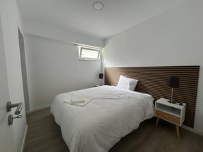 A bedroom with a bed, bedside table, lamp, and a small window. - Apartamento T2 com Terraço (Coimbra)