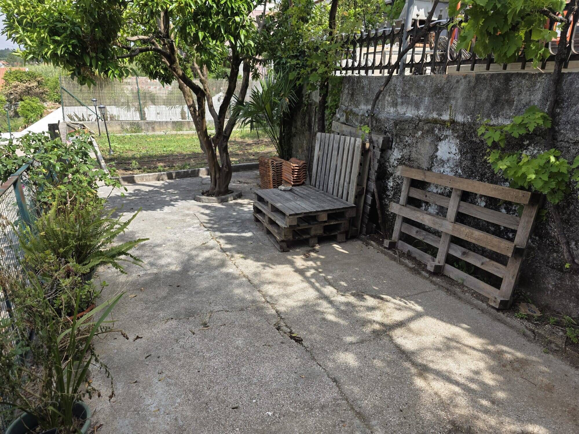 Un patio de concreto con un banco de madera, leña apilada y una cerca metálica.
