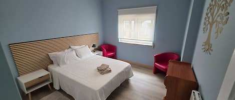 Un dormitorio con cama, mesitas de noche, escritorio, silla, radiador y ventana.