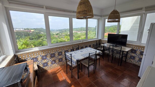 A dining area with a table and chairs, a television, and large windows with a view of greenery. - Quarto Suite Hope Vista Jardim (Coimbra)