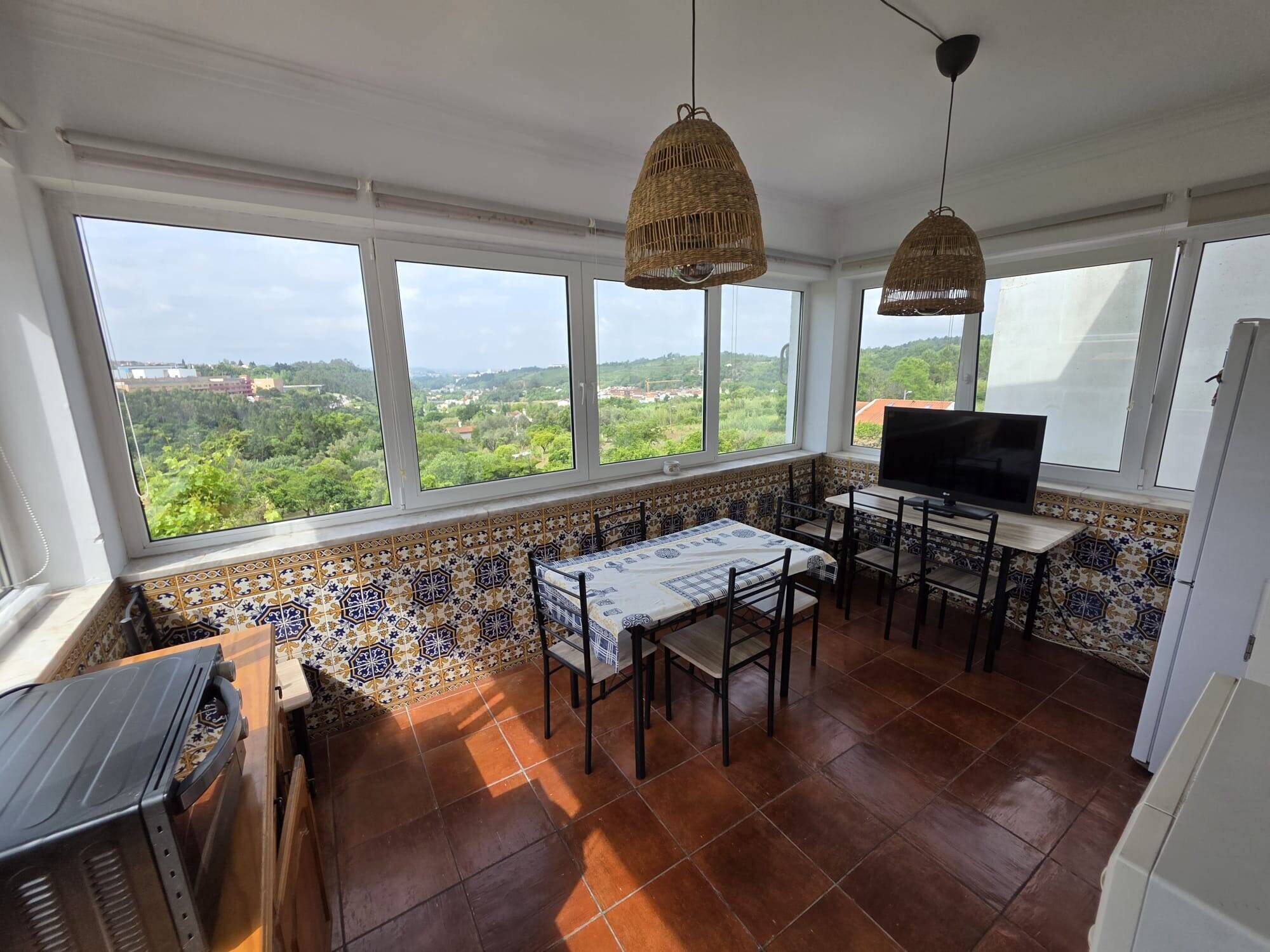 Un espace repas comprenant une table et des chaises, un téléviseur et de grandes fenêtres donnant sur un paysage verdoyant.