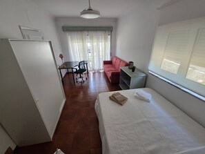 A bedroom with a bed, a desk, a chair, a sofa, and a window with blinds. - Quarto Sunrise com Varanda e WC (Coimbra)