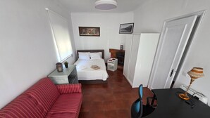 Habitación pequeña con cama, sofá rojo, una mesita y un escritorio con lámpara.