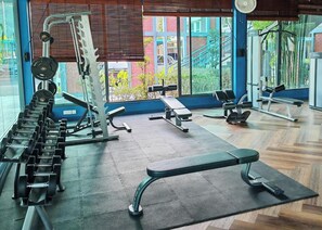 A gym with weightlifting equipment, benches, and a view of buildings through large windows.