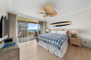 Ein Hotelzimmer mit Bett, Fernseher, Deckenventilator, Nachttisch mit Lampe und Blick auf einen Balkon mit Pool.