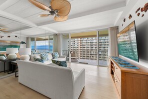 Een moderne woonkamer met een bank, een televisie, een plafondventilator en een balkon met uitzicht op een stadsbeeld.