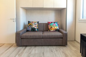 A brown leather sofa with colorful pillows, a wooden floor, and a window to the right. - HouSmart Greta Garbo 12 (Bologna)