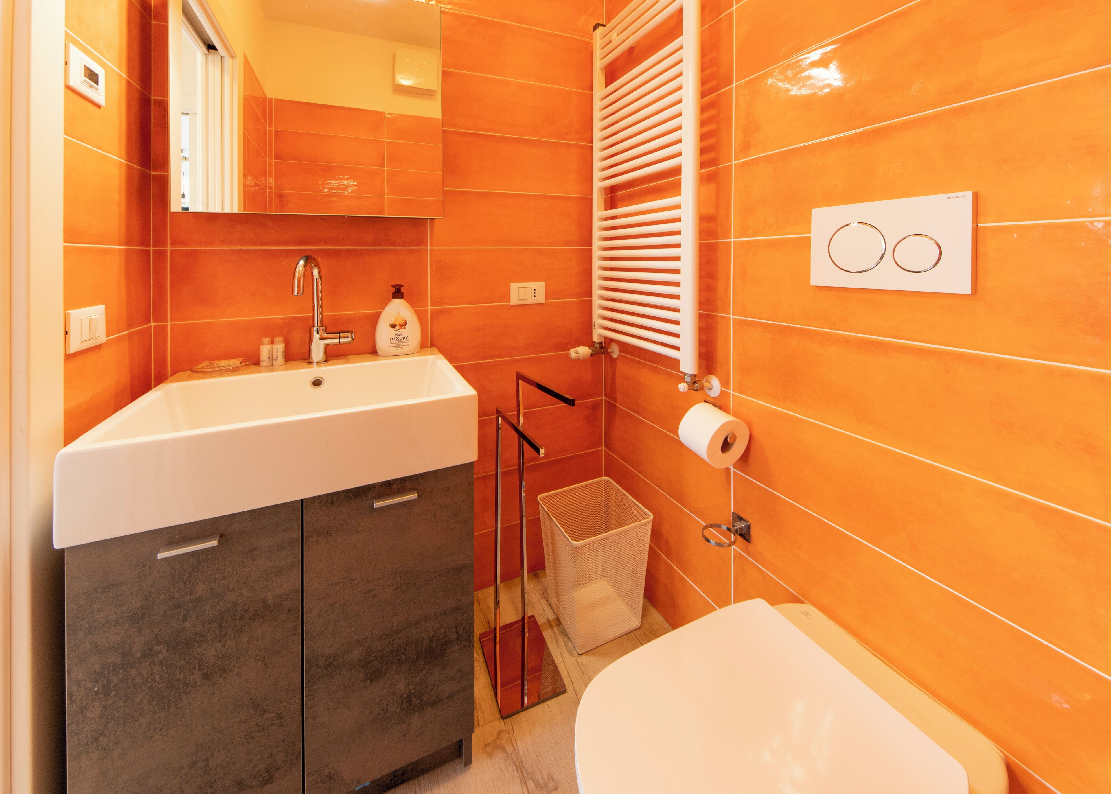 Une salle de bain avec un lavabo blanc, un miroir et des toilettes, des murs recouverts de carreaux orange et un porte-serviettes.