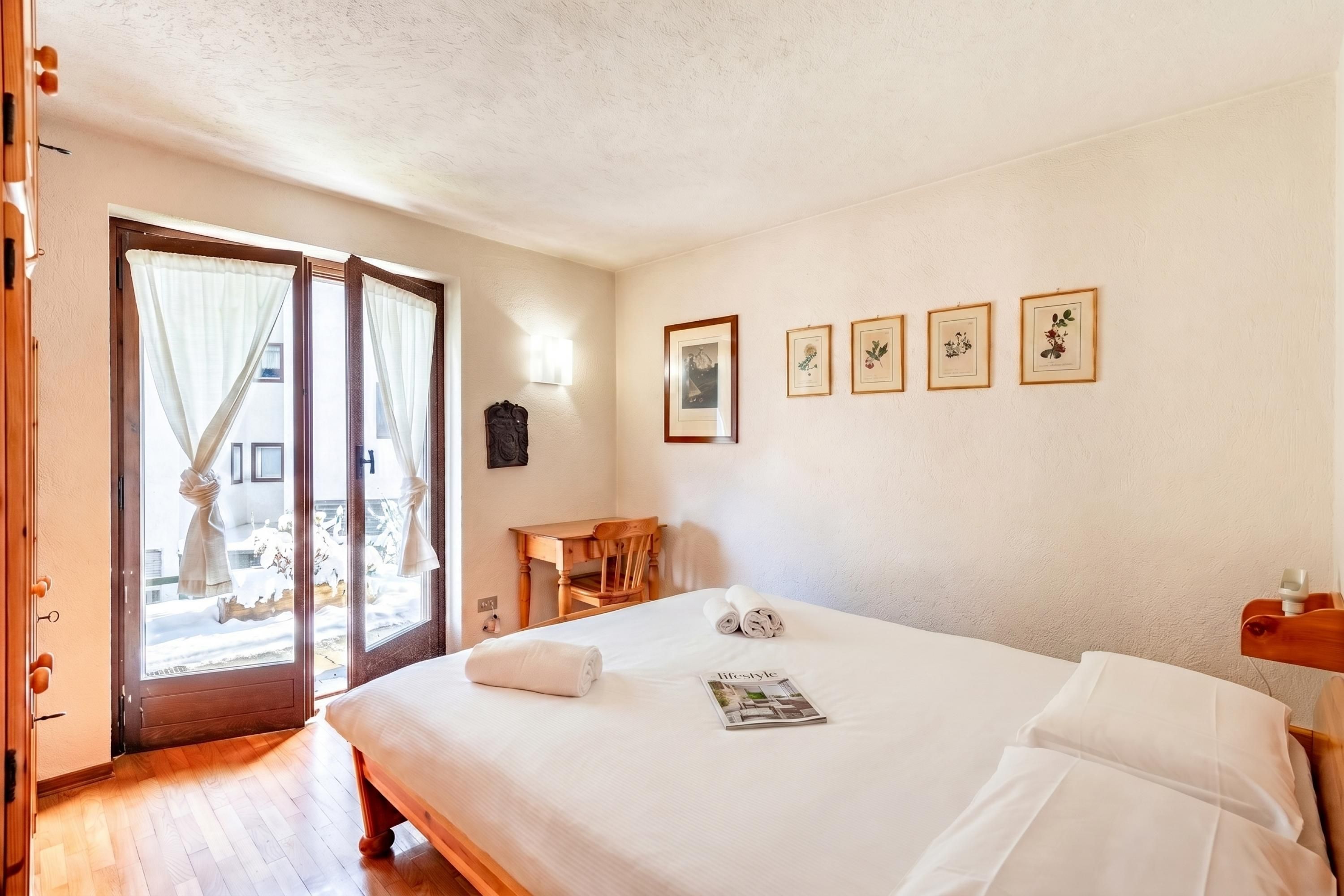 A hotel room with a bed, a desk, a chair, and a window with curtains. There are framed pictures on the wall.