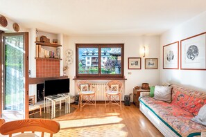 Un salon comprenant un canapé, deux fauteuils, un téléviseur et une fenêtre donnant sur des immeubles.