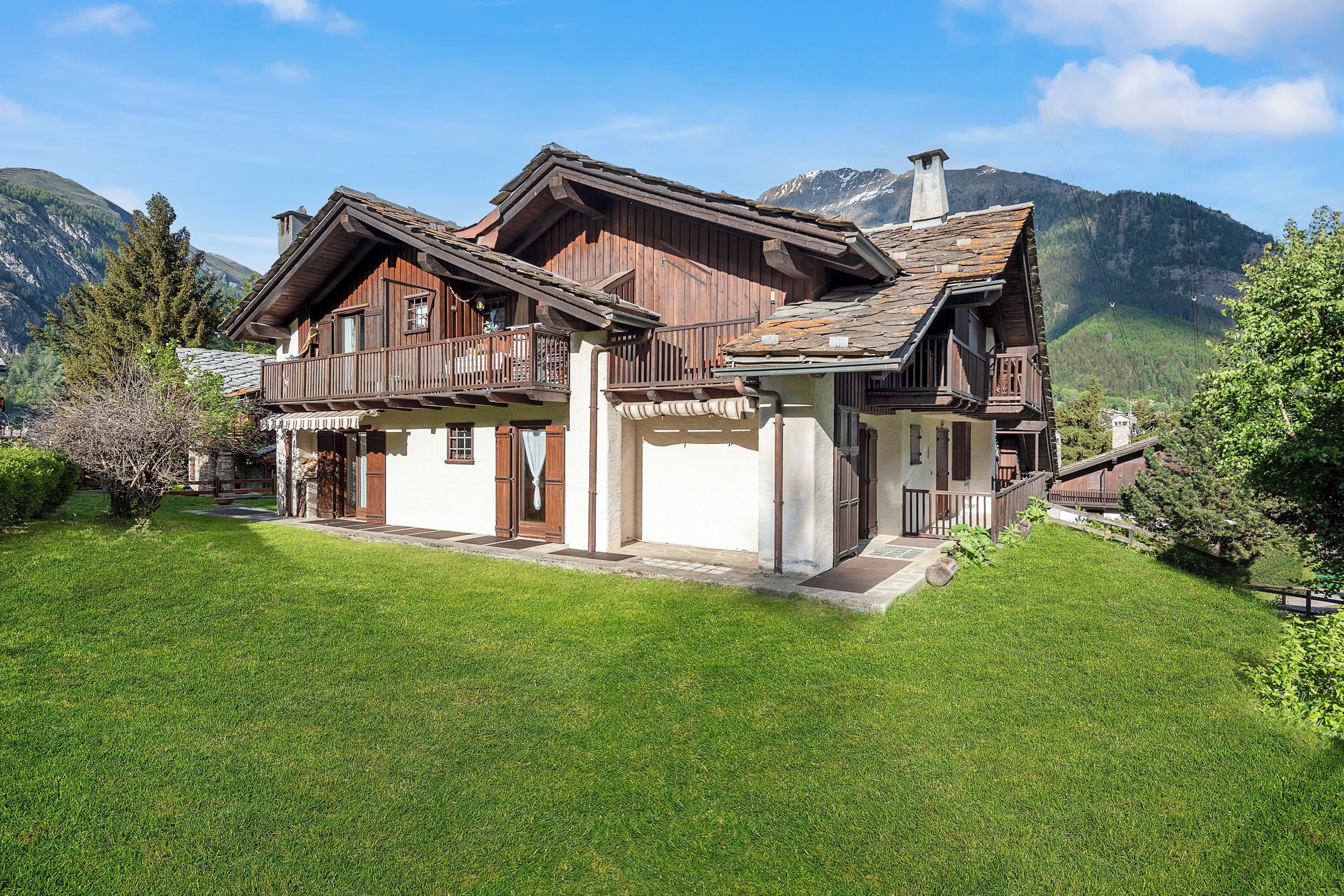 A two-story wooden chalet with a balcony, set against a mountainous backdrop.