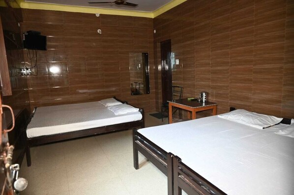 A hotel room with two single beds, a small table with a chair, a ceiling fan, and a TV mounted on the wall.