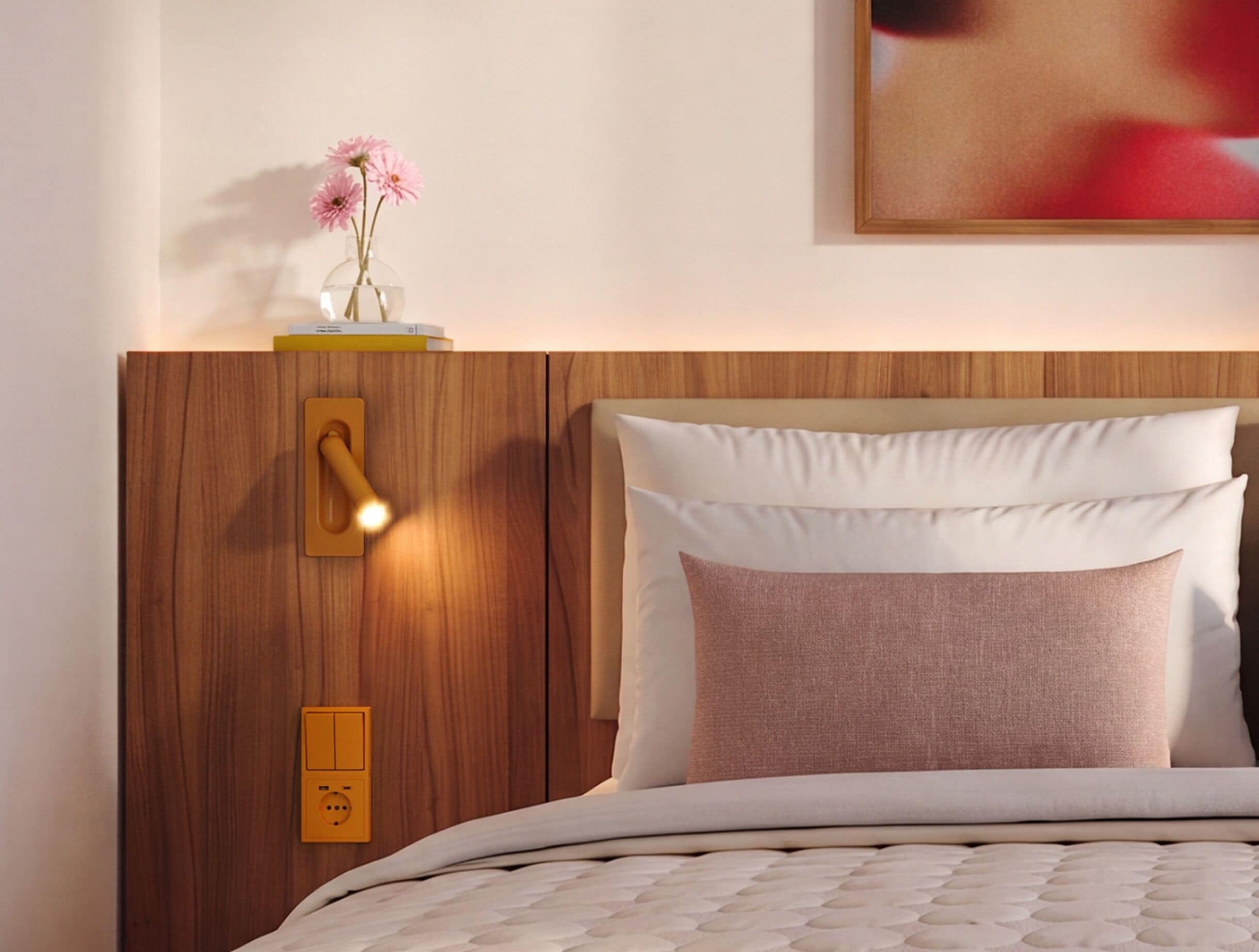 Cama bem arrumada com colcha branca texturizada, cabeceira de madeira, mesa de cabeceira com um vaso de flores cor-de-rosa e luminária fixada na parede.