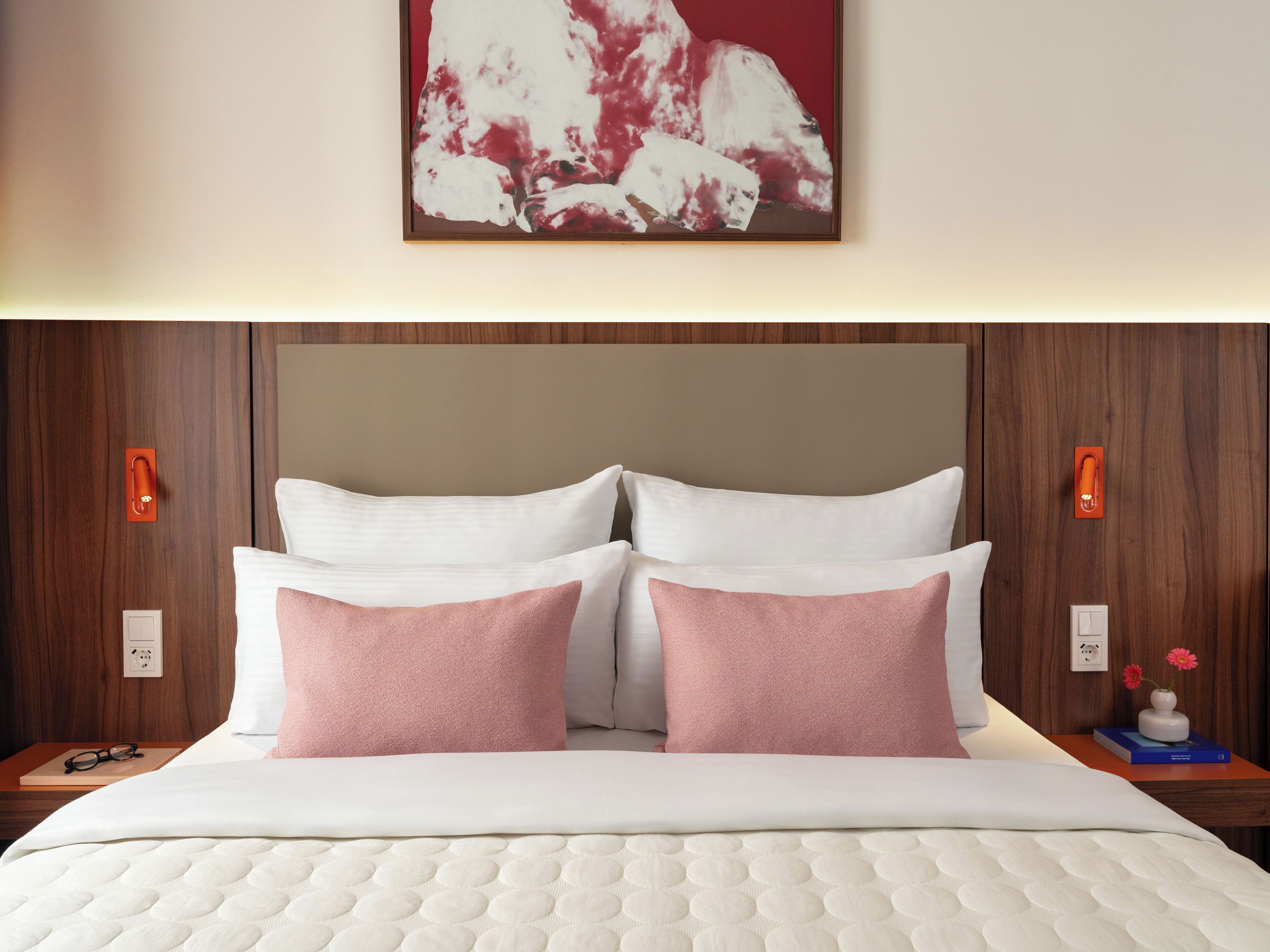 A hotel room with a bed, two pillows, a nightstand, a wall-mounted light, and a painting.