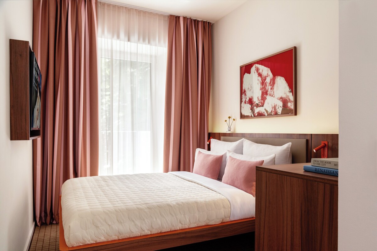 Een hotelkamer met een bed, een nachtkastje, een tv aan de muur, een schilderij en een raam met gordijnen.