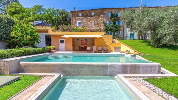 Una piscina con solarium, un edificio in pietra e vegetazione rigogliosa.