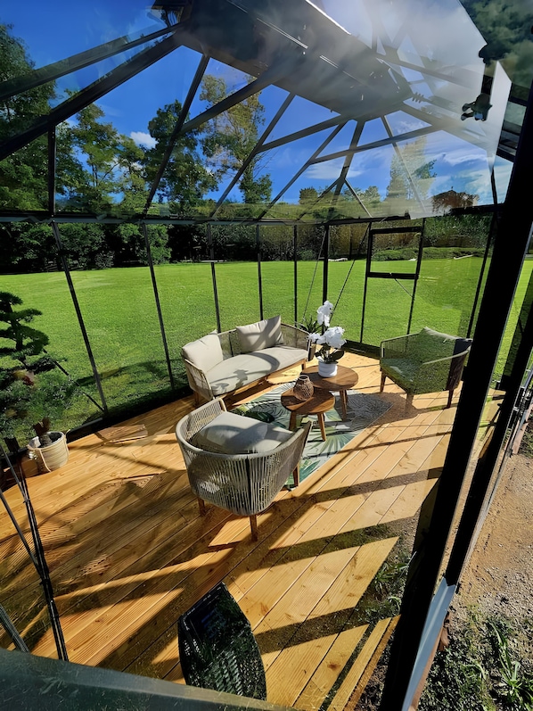 A covered outdoor seating area with a wooden deck, a cushioned chair, a small table, and a potted plant.