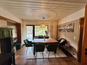 Uma sala de jantar com mesa de madeira, cadeiras verdes, luminária pendente e vista pela janela.