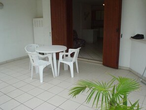 Un pequeño patio con una mesa y sillas blancas, una planta en maceta y una puerta que da a un espacio interior.