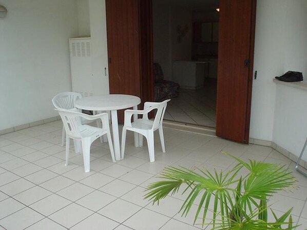 Une petite terrasse avec une table et des chaises blanches, un pot de fleurs et une porte donnant sur un espace intérieur.