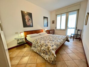 Een slaapkamer met een bed, een klein tafeltje, een stoel en een raam met gordijnen.