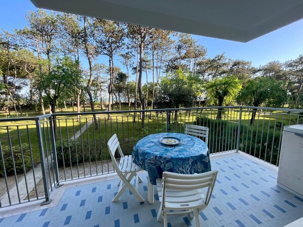 Een balkon met een tafel en stoelen, uitzicht op een park met bomen, en een betegelde vloer.