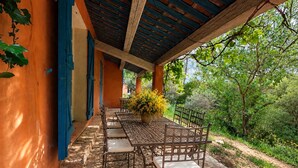 Un patio exterior con mesa y sillas, rodeado de follaje y una zona cubierta con contraventanas azules.