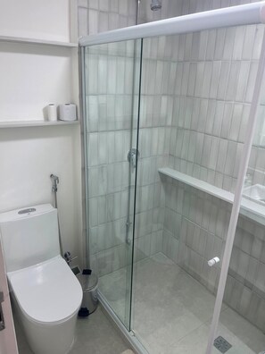 A bathroom with a glass-enclosed shower, a toilet, and a shelf with toiletries. - NEW Fully Equipped Apartment in Imbetiba (Macaé)