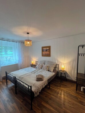 A bedroom with a bed, two bedside tables, a hanging light fixture, a window with curtains, and a painting on the wall. - Relaxation in the Eifel - nature, peace and comfort for families, couples and dog lovers (Seffern)