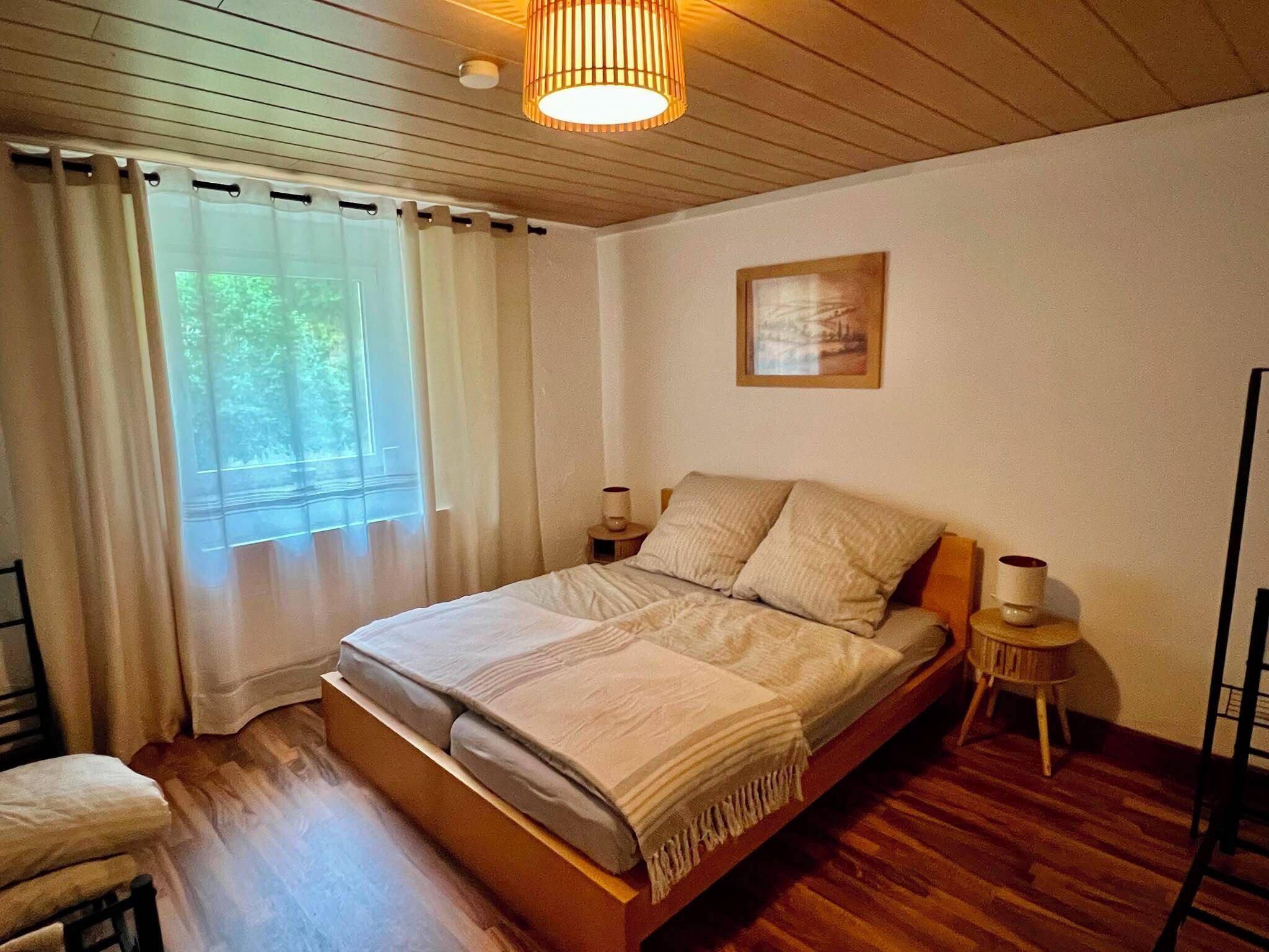 Un dormitorio con una cama de madera, una mesita de noche con lámpara, una ventana con cortinas y un cuadro en la pared.