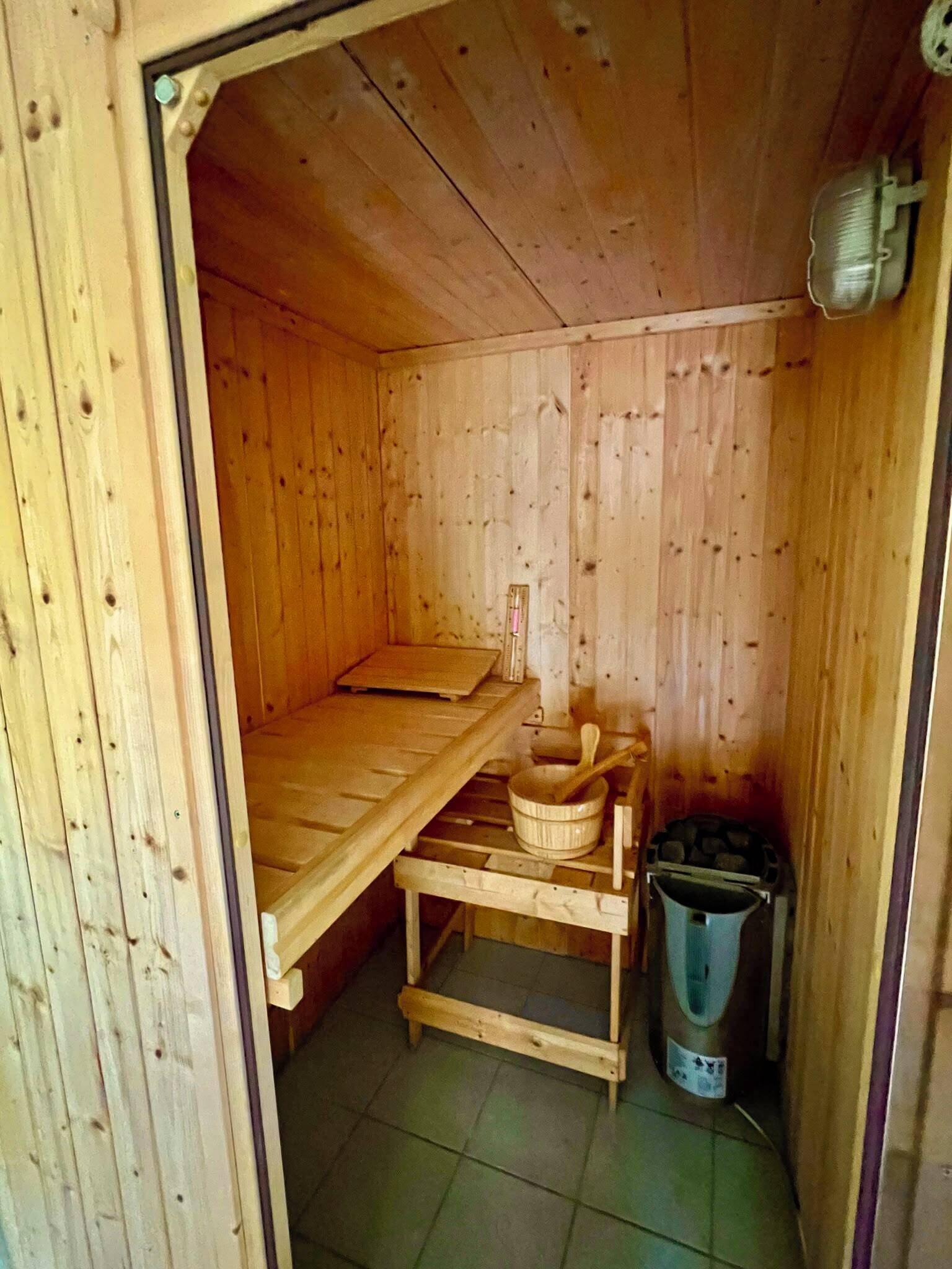 A wooden sauna room with a bench, a wooden table with a bowl and a ladle, and a trash bin.