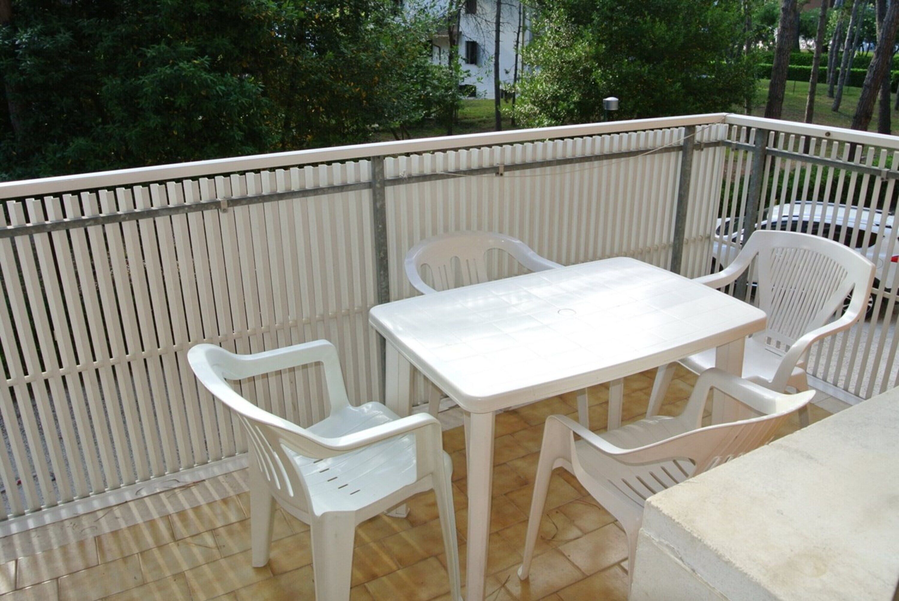 Un balcon avec une table et des chaises blanches, un sol carrelé et une vue sur les arbres et les bâtiments.