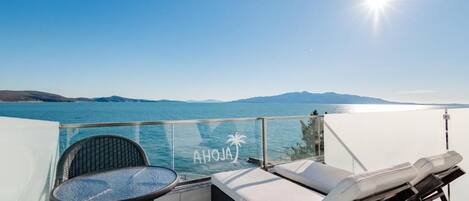 A balcony with a glass railing, a wicker chair, a lounge chair, and a view of the sea and mountains.
