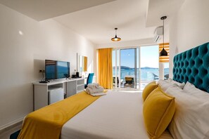A hotel room with a bed, a desk with a television, a chair, and a view of the outdoors. - Hotel Aloha (Sarande)