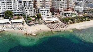 An aerial view of a coastal area with a beach, multiple buildings, and a clear blue sea. - Hotel Aloha (Sarande)
