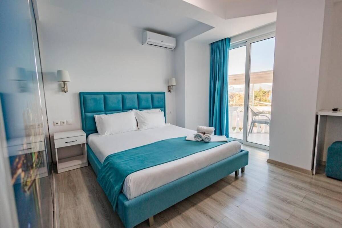 A modern hotel room with a large bed, a balcony with blue curtains, and a view of the outdoors.