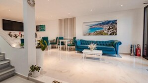 A modern living room with a blue sofa, a white coffee table, and a painting of a coastal landscape. - Hotel Aloha (Sarande)