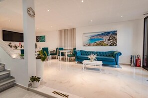 Uma sala de estar moderna com um sofá azul, uma mesa de centro branca e uma pintura de uma paisagem costeira.