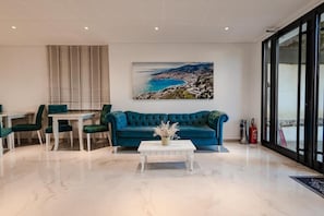 A modern living room with a teal sofa, white coffee table, and a painting of a coastal landscape. - Hotel Aloha (Sarande)