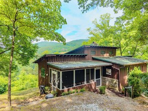 Un chalet rustique doté d’une véranda grillagée, entouré d’une végétation luxuriante et de collines.