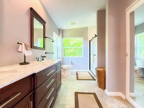Une salle de bain dotée d’un grand miroir, de meubles de rangement aux couleurs sombres, d’un plan de toilette en marbre et d’une fenêtre donnant sur un paysage verdoyant.