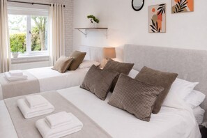 A hotel room with two beds, a window with curtains, a bedside table with a lamp, and wall art. - Modern coastal retreat – 5 bed home offering space and style for up to 12 guests (Folkestone)