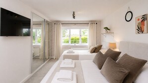 A modern hotel room with a large bed, a smaller bed, a TV, a window with curtains, and a plant on a shelf. - Modern coastal retreat – 5 bed home offering space and style for up to 12 guests (Folkestone)